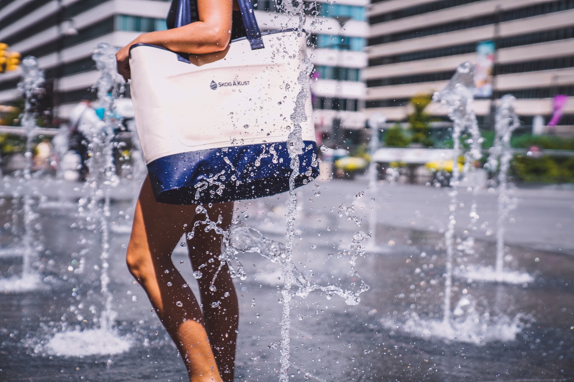 The Skog A Kust waterproof ToteSak Tote Bag being used by a women to protect their belongings while walking through a spouting water feature in the city